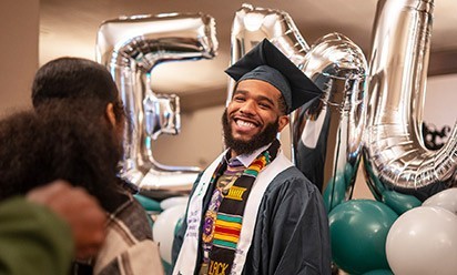 A student in cap and gown smiles in front of silvery EMU balloons