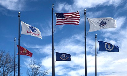 Flags representing U.S. military branches fly against a blue sky.