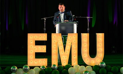 Scott Wetherbee on the stage amid green and white balloons and a lighted EMU sign.