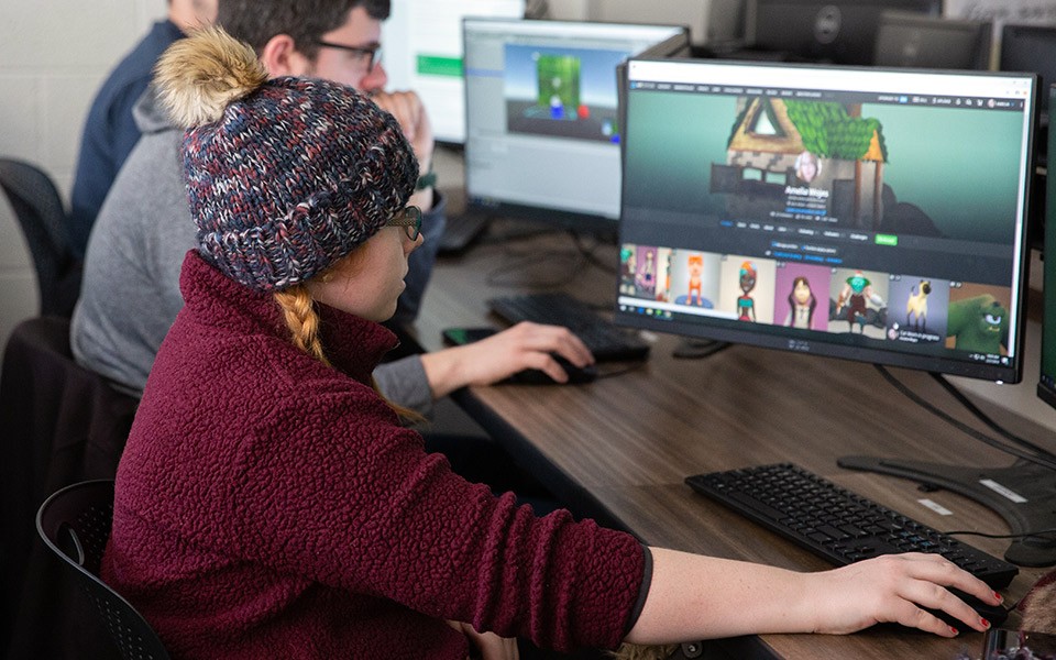 A student wearing a winter hat works on a colorful animation project on a classroom computer with other students and computers in the background.