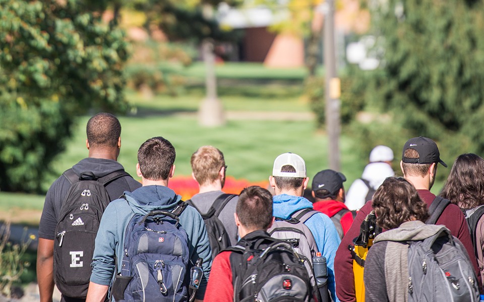 Students, seen from the back, walk on campus with their backpacks on.