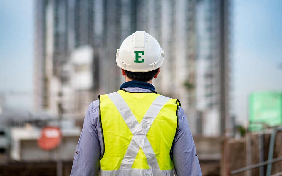 A person seen from behind at a construction site wears a white hard hat with a green block E on it