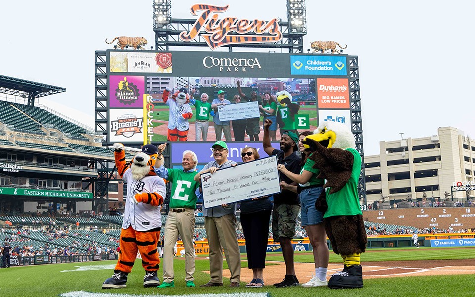 Paws, Walter Kraft, President James Smith, Connie Ruhl-Smith, Alex Simpson, Ameera Salman, and Swoop hold a big check for the EMU Student Emergency Fund at TRUEMU Night at Comerica Park.