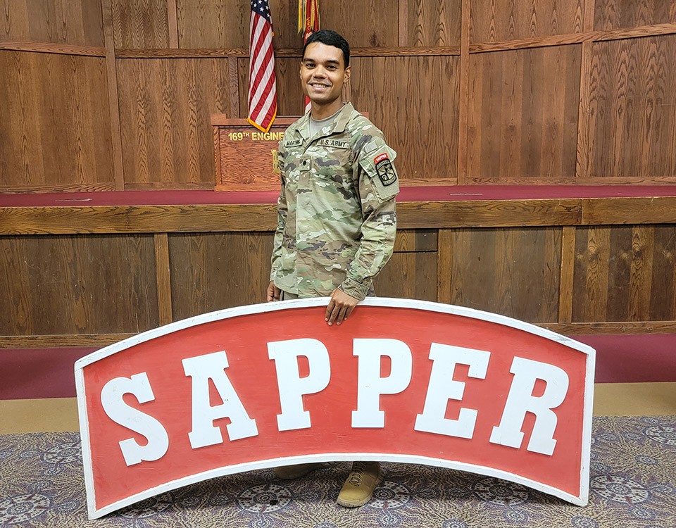 Cadet Renan Martins in his military uniform stands behind a large Sapper sign