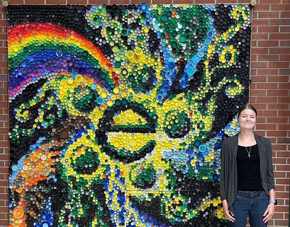 Olivia Robinson stands in front of the large, colorful mosaic she created using different bottle caps.