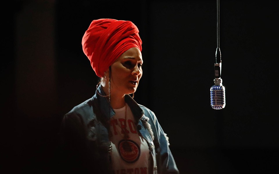 Mama Sōl, with her hair wrapped in a bright red scarf, stands by a microphone hanging from its cord.