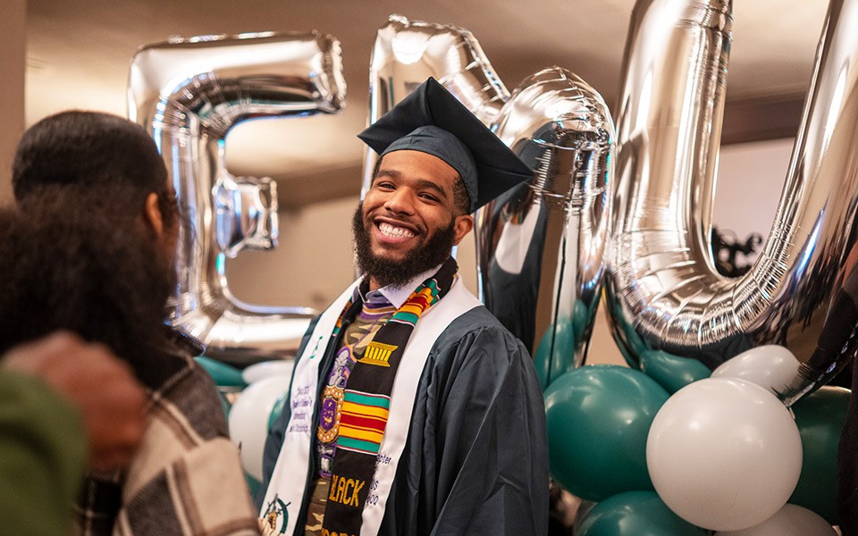 A student of color smiles in his cap and gown in front of green, white and silver EMU balloons.
