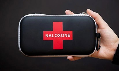 a hand holds a black Naloxone kit with a red cross on it