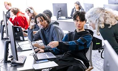 Students at computers talk with a professor in the classroom.