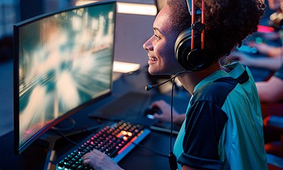 A young black woman is smiling and wearing headphones while playing a video game in an esports setting with computers around.