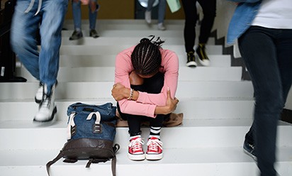 A student wearing a pink shirt and shoes sits on steps with their head down while other students walk past