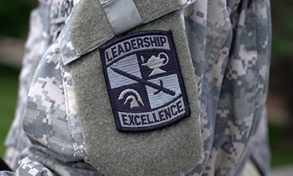 A camouflage uniform sleeve sports a patch embroidered with military symbols and the words "Leadership," and "Excellence."