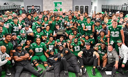 large group of football players in dark green jerseys gathered around head coach Chris Creighton, who is holding a trophy.