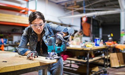 woman wearing jeans, safety glasses, and protective gloves, uses a power tool in the shop.