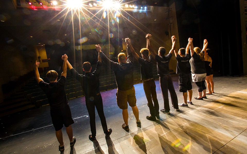 A group of EMU Theatre students hold hands and take a bow in a view from behind them on stage.