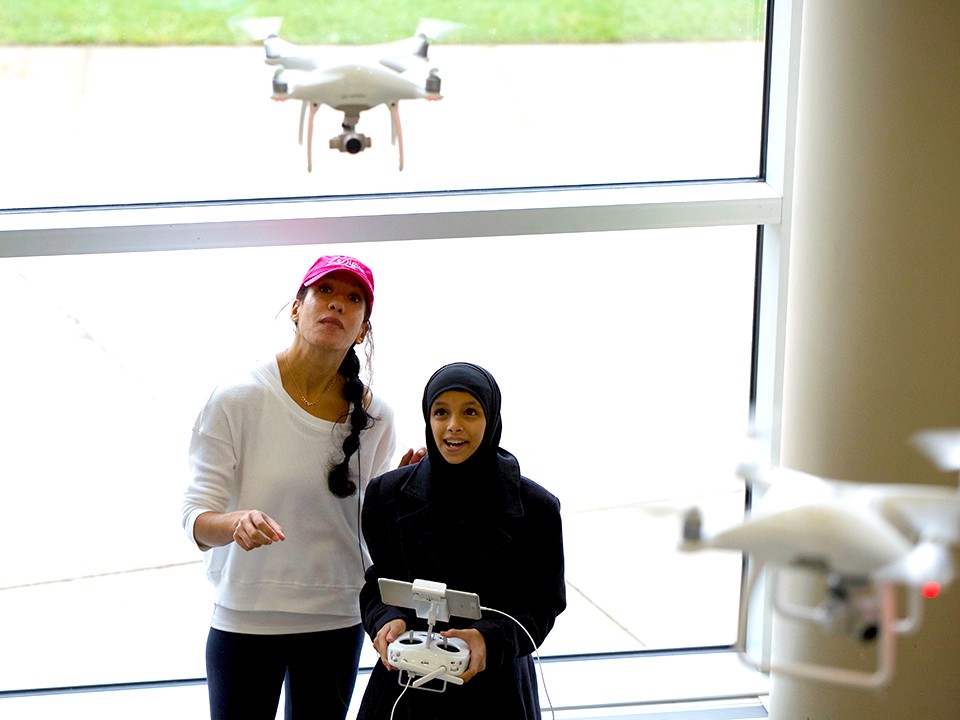 Kyla Berry and a young girl with a controller look up at a drone that is airborne at the Digital Divas event at the Student Center.