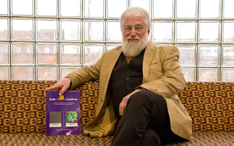 Joe Radding sits on a couch at the College of Business next to a copy of his book, "Suits Plus Creatives."