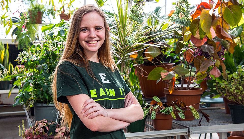 A student wearing an EMU t-shirt stands smiling in the greenhouse filled with plants.