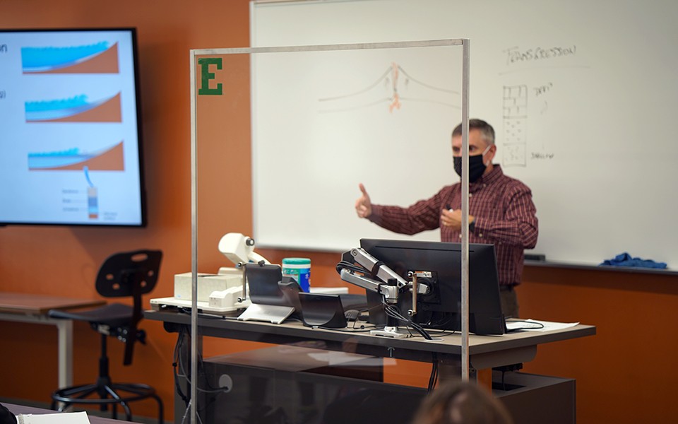 EMU Professor teaching an in-person class behind Plexiglas barrier.