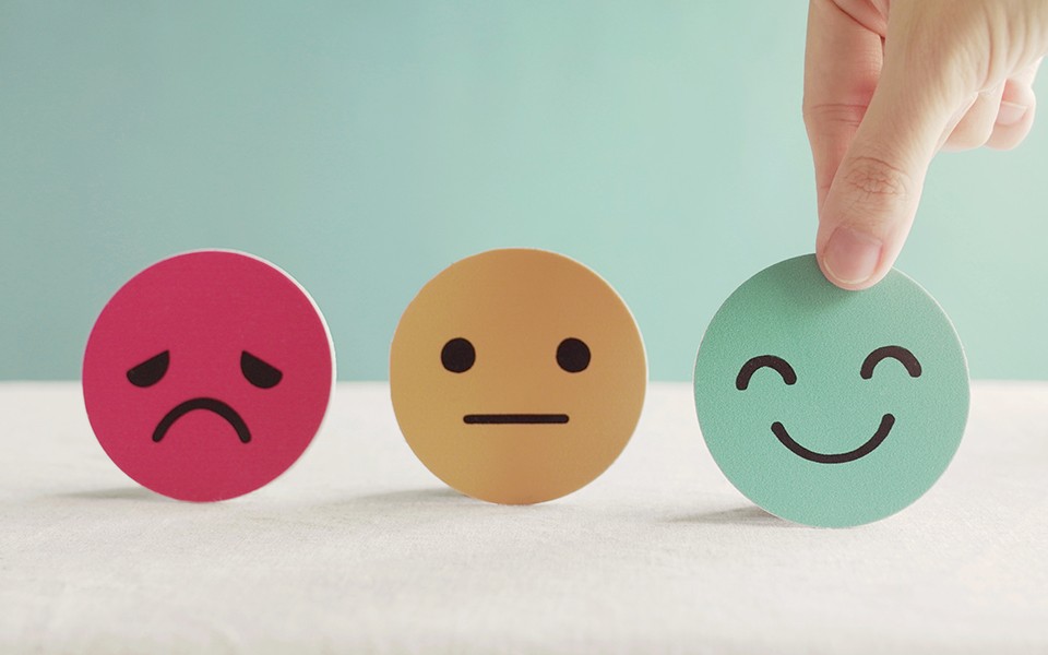 photo illustration with 3D emoji faces: sad, neutral and a person's hand holding the happy face