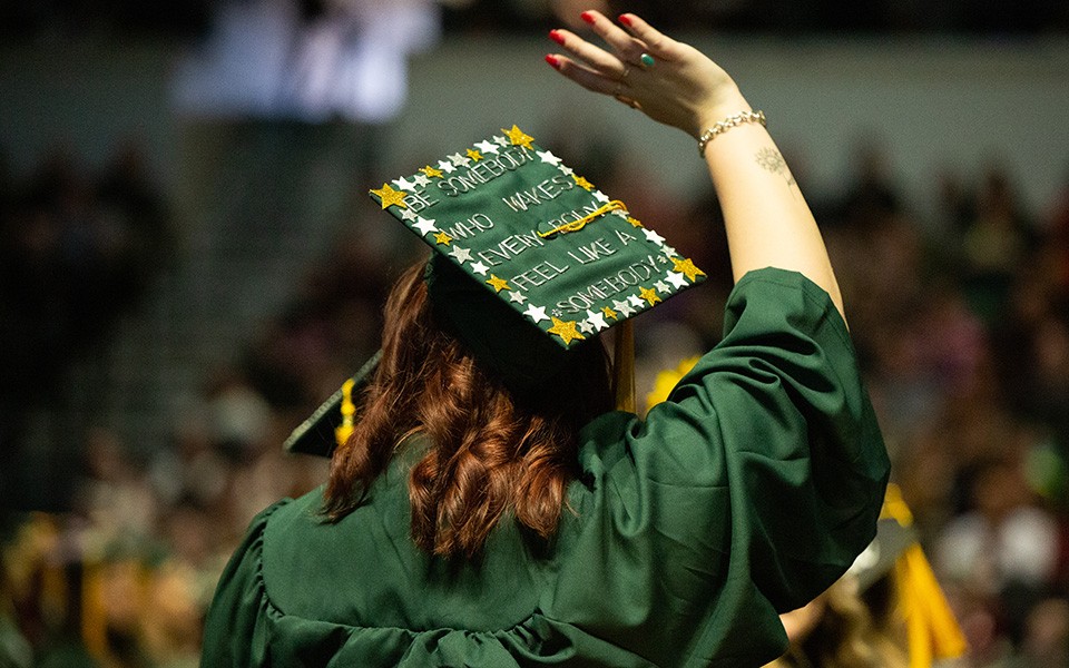 A grad wearing a mortarboard that says, "Be somebody who makes everybody feel like a somebody" waves to the crowd at commencement.