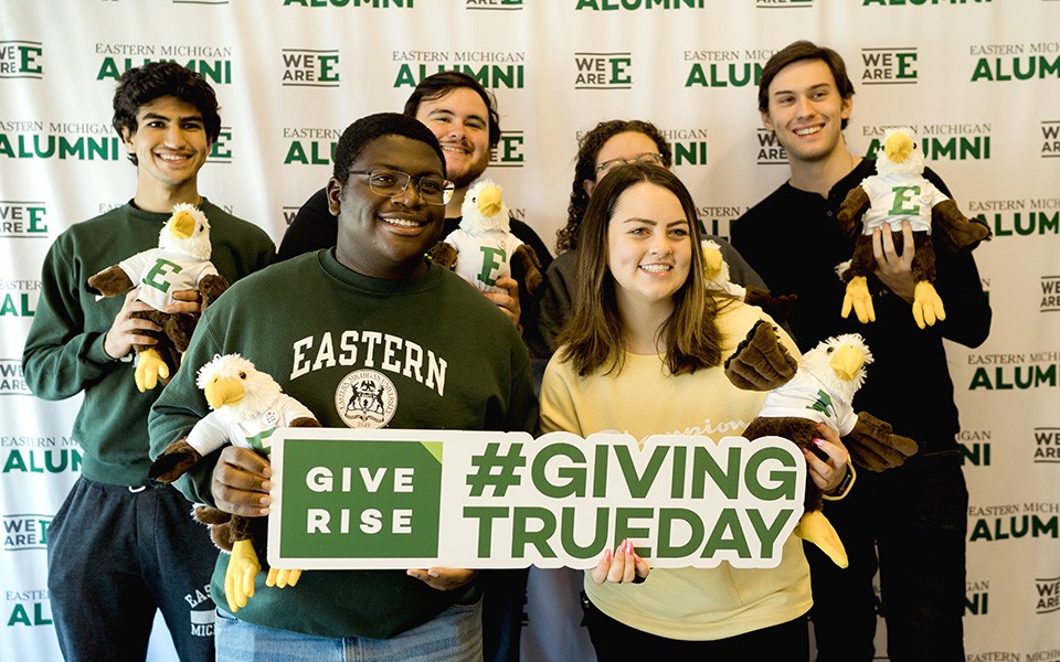 A group of students pose with stuffed Swoop eagles and a #GivingTRUEday sign in front of the Alumni Association backdrop.
