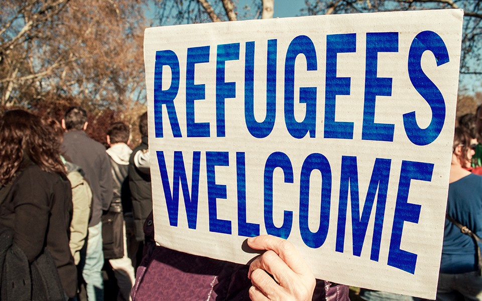 A person holds up a sign that reads, "Refugees welcome" in a group of people gathered outside.