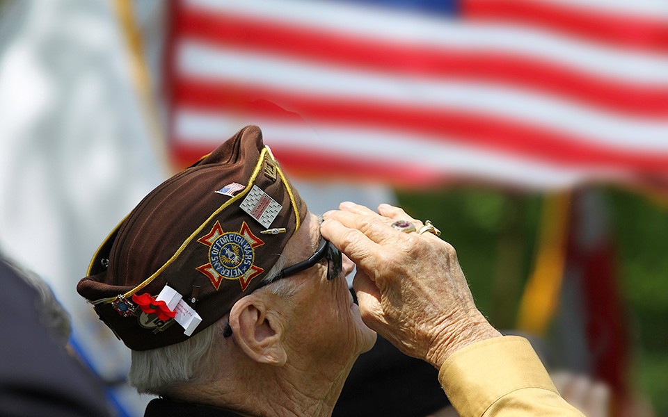 Stock photo of a gray-haired man in a hat decorated with a Veteran of Foreign Wars patch while saluting the American flag.