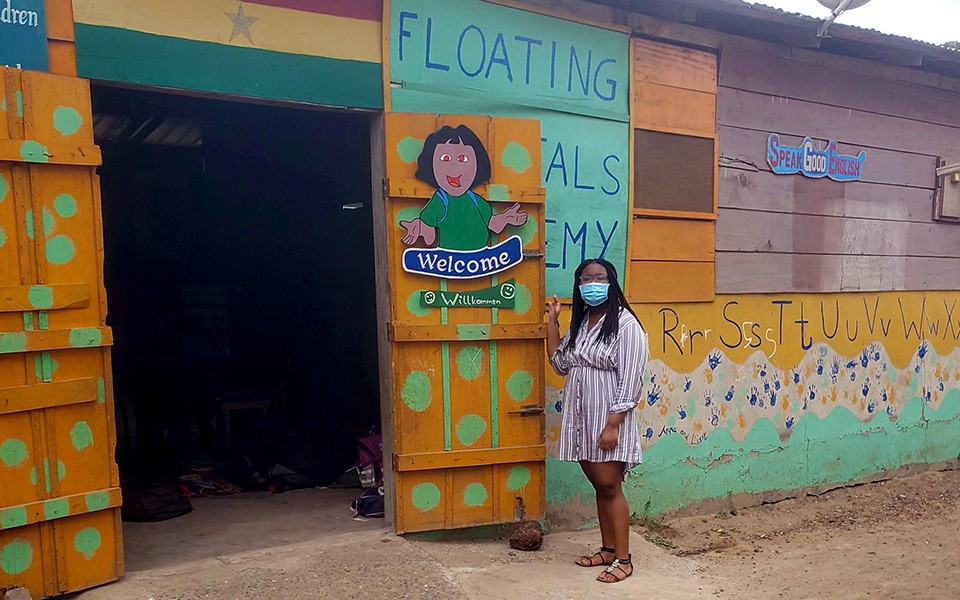 Cheyanne Roy, wearing a mask, stands outside the open door of the colorfully-painted Floating Crystals Academy school in Ghana.