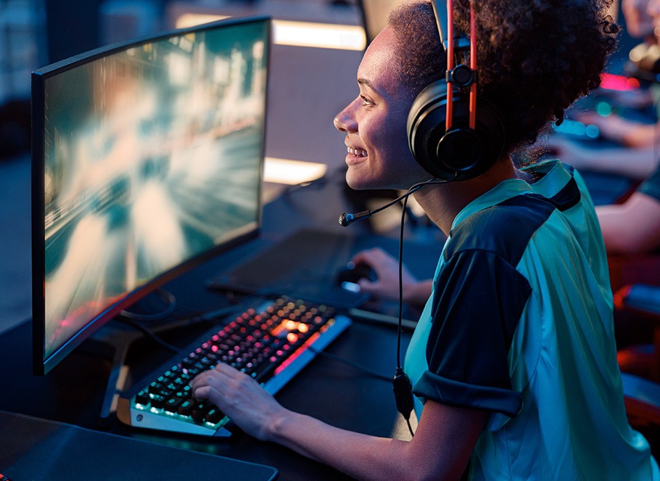 A young black woman is wearing headphones while playing a video game in an esports setting with many computer terminals all around.