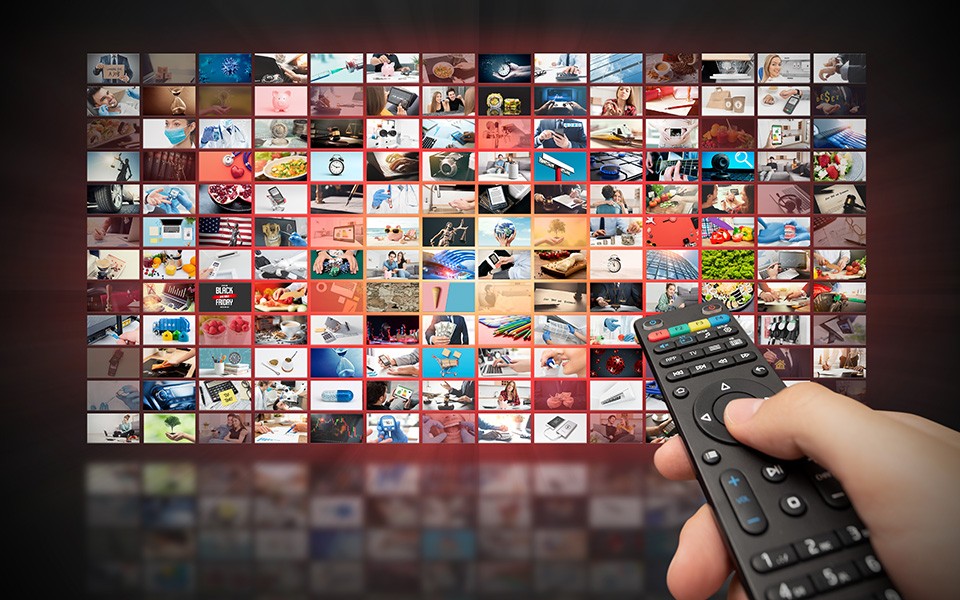 A hand is pointing a remote control at a large-screen TV that has a grid of small ad images on it.