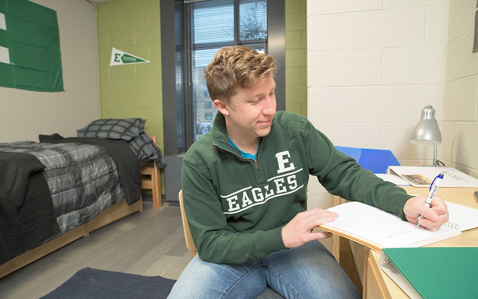 A student in an Eastern Eagles sweatshirt writes at the desk in his dorm room which is decorated with an EMU pennant and flag.
