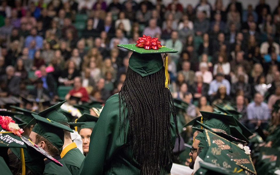 A graduate facing the crowd at the commencement ceremony with a bow on her mortarboard.