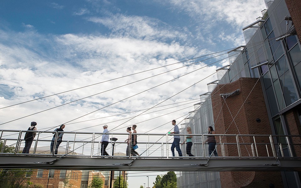 People walk across the bridge at the Mark Jefferson Science Complex on a partly cloudy day