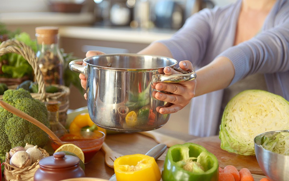 A woman's hands hold out a shiny cooking pot on a counter full of produce.