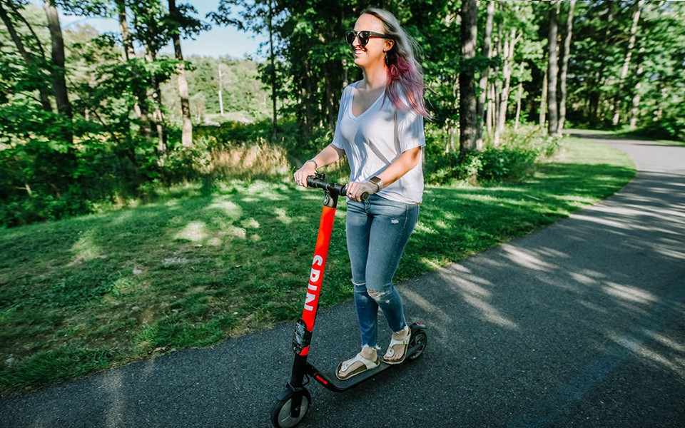 A young woman rides a Spin electric scooter on a tree-lined path.