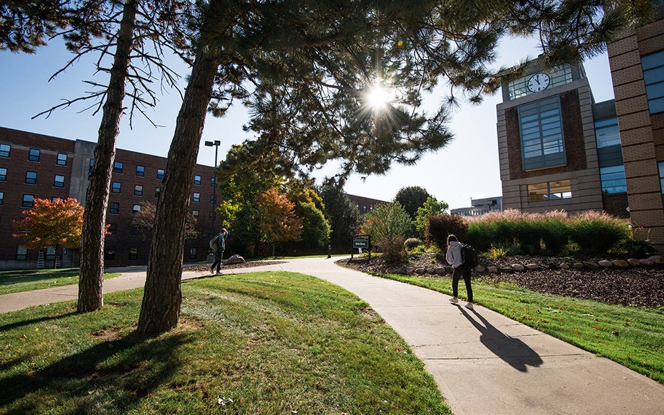 Students walk on campus by Halle Library on a fall day, with a burst of sunlight shining through the pines.