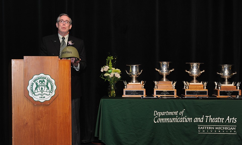 Dennis Beagen holds an Army helmet with Patton stenciled and stands at a podium next to a table of Forensics trophies at a past event.