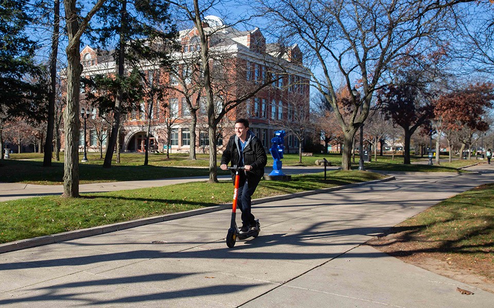 A student smiles while riding a Spin electric scooter across campus on a crisp fall day.