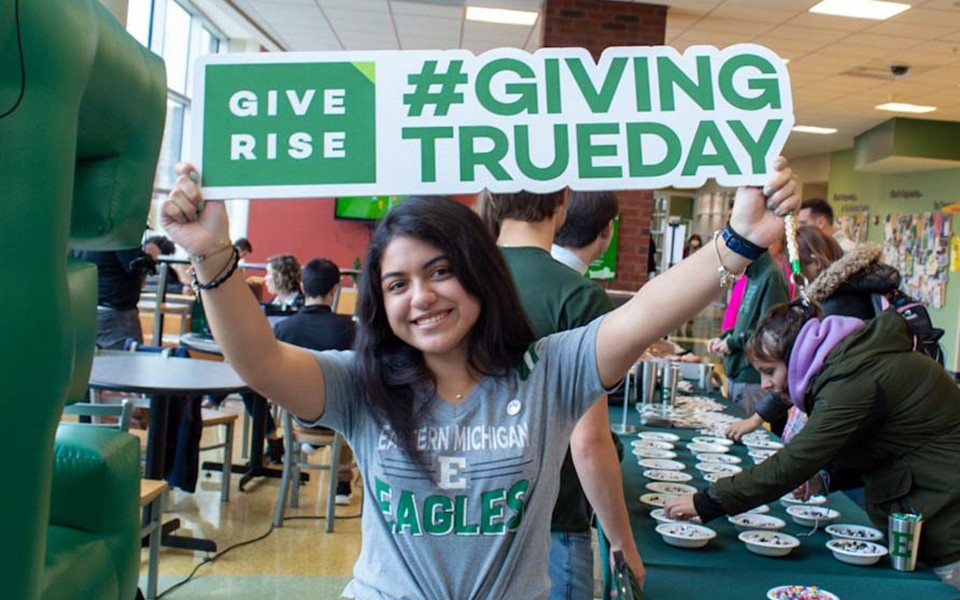 EMU student wearing an Eastern t-shirt holds up a Give Rise #GivingTrueday sign at the Student Center tables