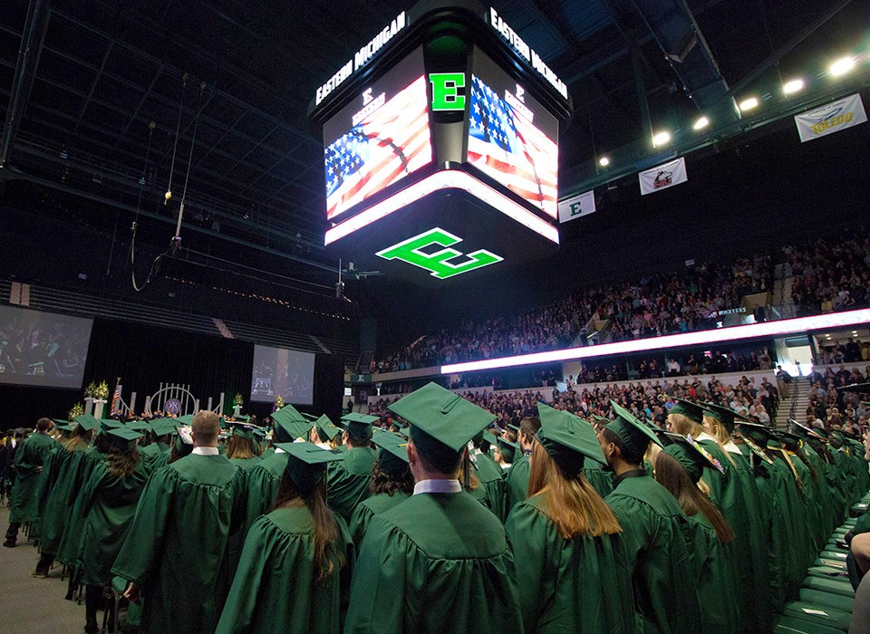Graduates in caps and gowns gathered in rows at the George Gervin GameAbove center under the big Block E and American flag on the scoreboard.