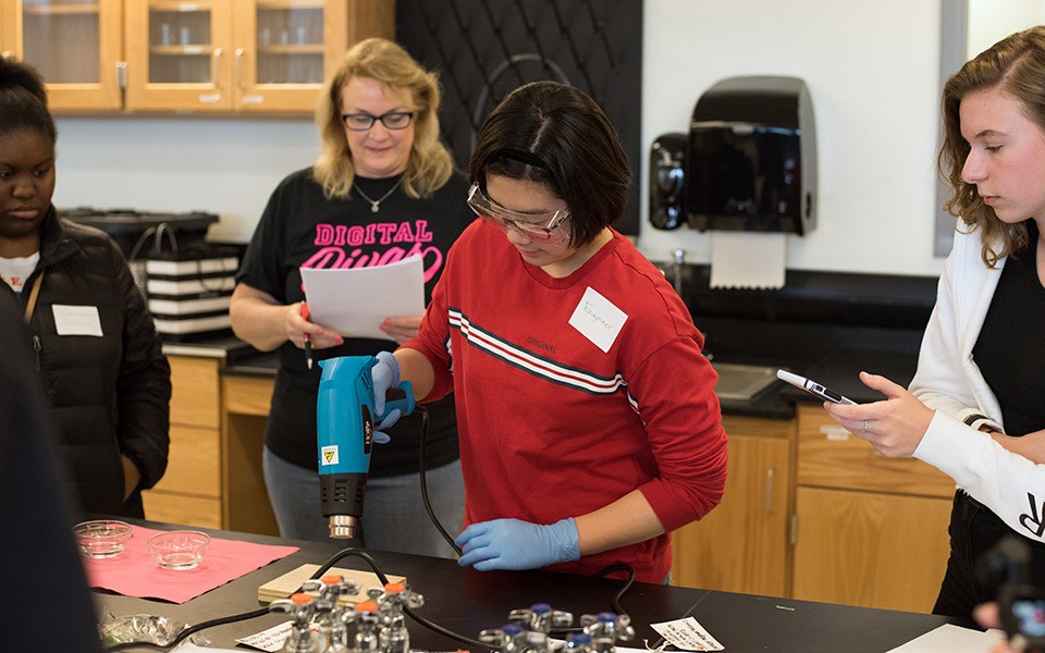 A young woman in a red shirt wears a name tag that says "Engineer," works on a project while an instructor from the Digital Divas event and other young women look on.