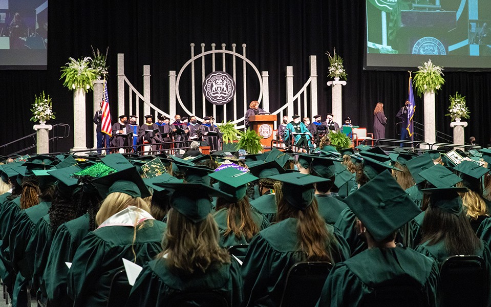 A view of the commencement platform with a speaker at the podium and chairs filled with grads in caps and gowns
