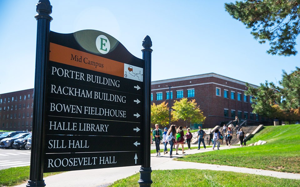 Mid-campus directional sign with students walking to class on a sunny day in the background