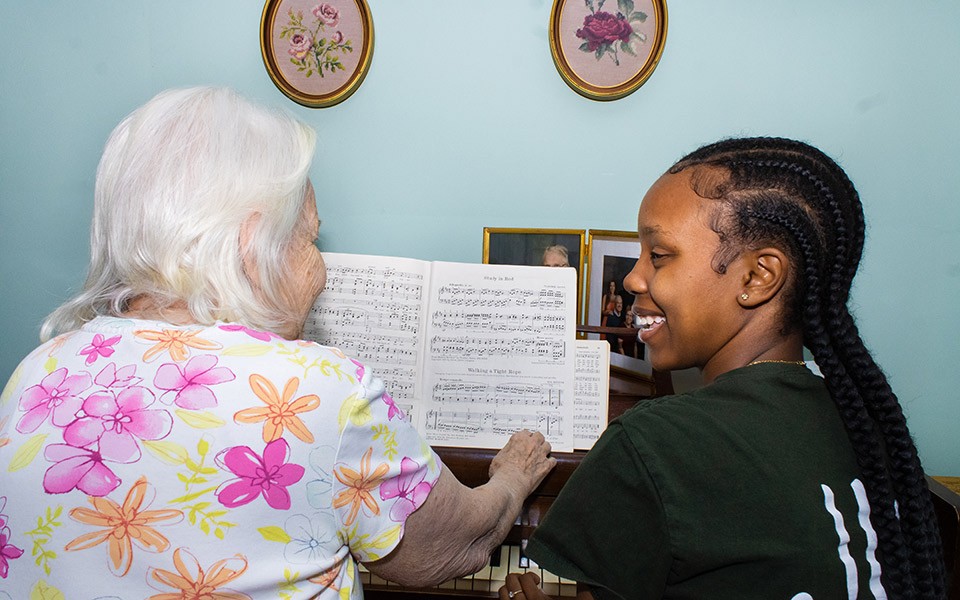 older adult with a young Loris hands volunteer look at music at a piano