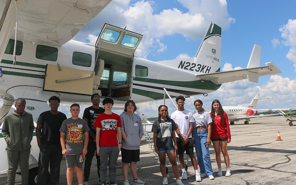A group of young people stand next to the airplanes outside at Willow Run Airport.
