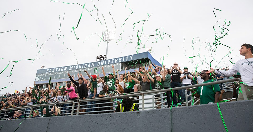 Fans in the stands at Rynearson Stadium cheer and toss green and white streamers and confetti in the air at a homecoming football game.