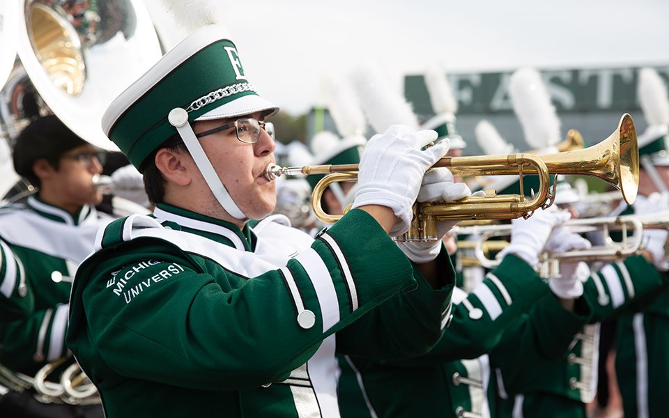 Trumpet player in EMU Marching Band uniform with more band members in the background.