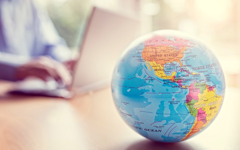 A globe sits on a table with a person on a laptop in the background.