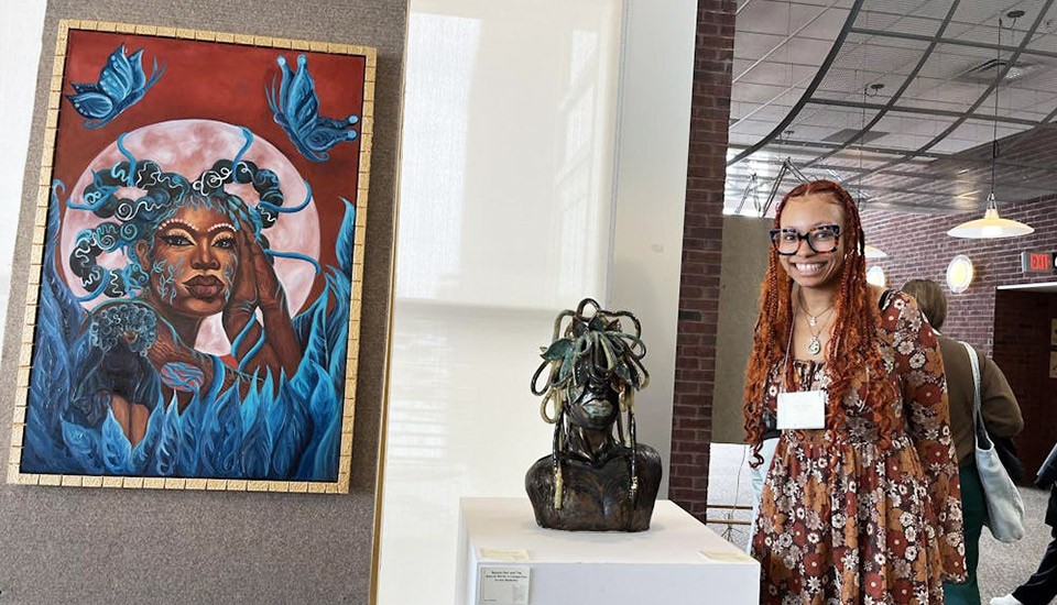 Nia Crutcher stands next to a sculpture and a painting that she's created and is exhibiting in her solo exhibit.